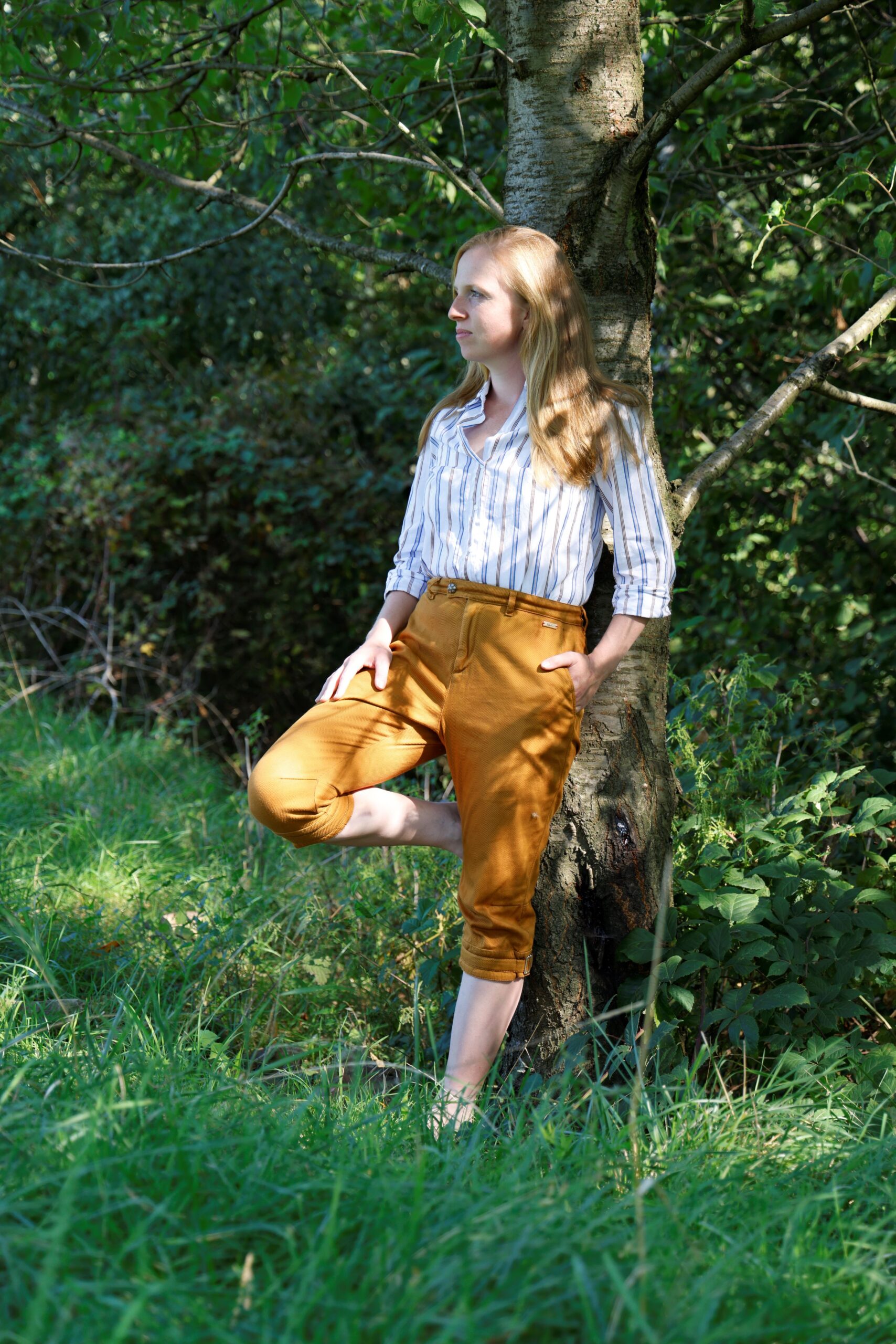 Coachin lehnt entspannt an Baum mit Hand in der Tasche und Fuß angelehnt in der Sicherheit, Klarheit und nachhaltige Lösungen für Macher zu finden